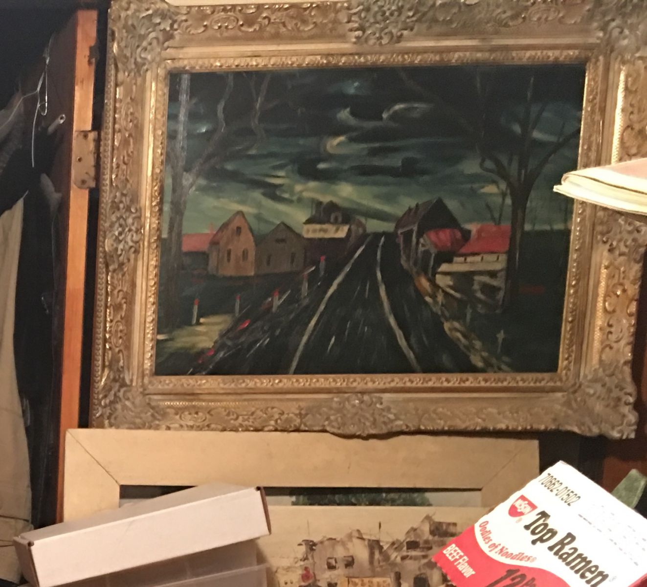 A moody framed landscape oil on canvas painting sits atop other works of art and a box of ramen noodle soup inside this estate.